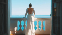 Mariée de dos en robe de dentelle à manches longues et dos nu se tenant sur un balcon avec balustrade blanche ornée, vue panoramique sur la mer Méditerranée bleue turquoise