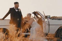 Couple de mariés posant près d'une voiture décapotable blanche vintage dans un champ de blé doré au coucher du soleil en Grèce