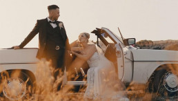 Bride and groom pose beside a vintage white convertible car in a golden wheat field at sunset in Greece.