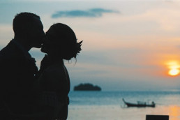 Silhouette d'un couple de mariés s'embrassant au coucher du soleil devant la mer, avec un bateau de pêche et une petite île visible
