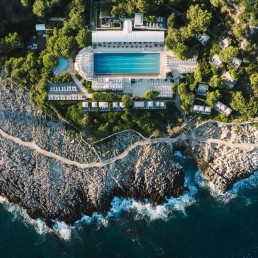 Spectacular aerial view of the Grand Hôtel du Cap Ferrat, with its turquoise Olympic-size swimming pool surrounded by white deckchairs, rocky cliffs and deep-blue Mediterranean Sea.