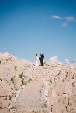 Bride and groom standing at the top of an ancient stone staircase on a rocky hillside under a blue sky with white clouds.