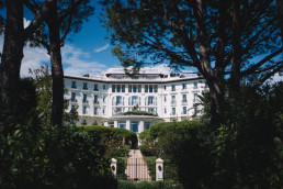 Façade blanche du Grand Hôtel du Cap Ferrat encadrée par des pins parasols méditerranéens, avec portail d'entrée et jardins luxuriants