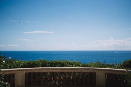 Panoramic view of the deep-blue Mediterranean Sea, with a white balustraded terrace in the foreground and vegetation.