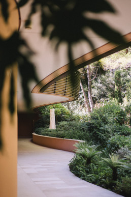 View from inside a covered terrace overlooking a lush Mediterranean garden with tropical vegetation, palm trees and paved driveway
