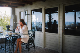Mariée assise sur une chaise en fer forgé sur une terrasse couverte avec colonnes blanches, vue panoramique sur la mer en arrière-plan