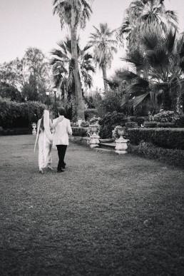 Photo noir et blanc de couple de mariés marchant main dans la main sur pelouse d'un jardin sicilien avec grands palmiers et fontaines en pierre