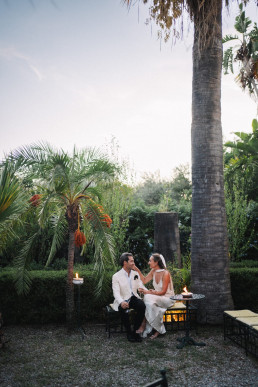 Couple assis sous palmier au crépuscule dans jardin sicilien avec bougies et ambiance romantique