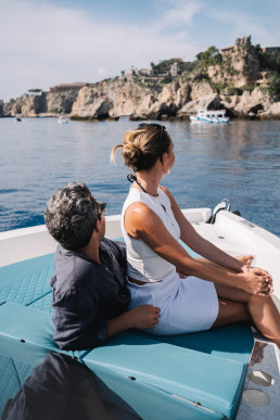 Couple sitting on boat admiring Sicilian coast with cliffs and medieval castle in background