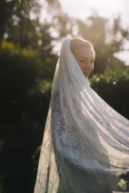 Smiling bride wrapped in her long white lace veil, fluttering in the wind, photographed against a golden backlight at sunset in a garden.
