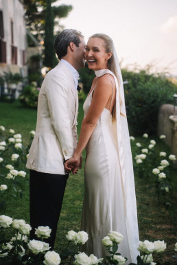 Bridegroom tenderly kissing the cheek of his smiling blonde bride in a white halter dress, hand in hand in a garden with white roses.