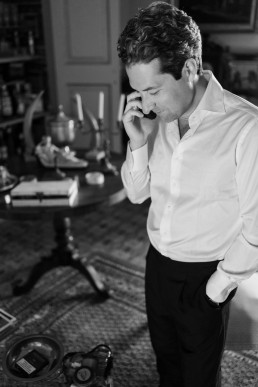 Black and white portrait of an elegant groom in white shirt and black pants talking on the phone in a luxurious living room with leather armchairs and coffee table.