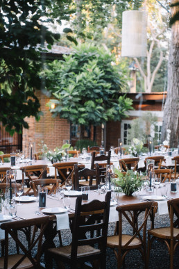 Tables de réception dressées en plein air sous des arbres, avec nappes blanches, chaises en bois et décoration florale