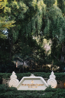 Ornamental white Four Seasons Resort Chiang Mai entrance sign with gold lettering, framed by verdant tropical trees and a trimmed hedge