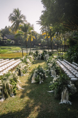Outdoor ceremony aisle at the Four Seasons Chiang Mai with white floral arch, white chairs lined up, beige fabric garlands and rice paddies in the background with palm trees.