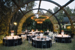 Spectacular outdoor wedding reception at night with large arches illuminated with gold string lights, round black tables with white tablecloths, transparent chairs and hanging chandeliers.