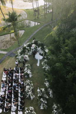 Aerial view of outdoor wedding ceremony with white floral arch near rice paddies and palm trees