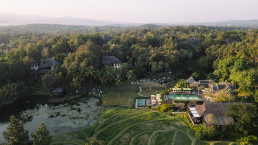 Panoramic aerial view of a tropical resort at Four Seasons Chiang Mai, with green rice terraces, water basin, palm trees, traditional Thai villas and misty mountains in the background.