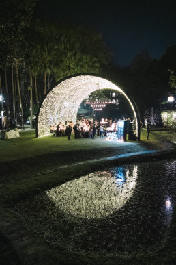 Night-time wedding reception under an illuminated stone arch, reflected in a pool of water, with trees in the background.