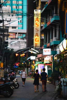 Busy street in Chiang Mai, Thailand, at dusk with colorful illuminated massage signs, electric cables, scooters and pedestrians, typical urban atmosphere.