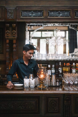 Barman in blue shirt working behind a traditional Thai carved wooden bar with liquor bottles, crystal glasses and decorative antique mirror