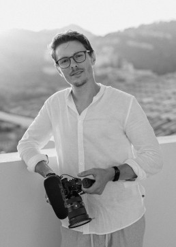 Black and white portrait of a smiling videographer with glasses and white shirt holding a professional camera, blurred mountains in the background.