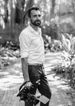 Black and white portrait of a bearded videographer in a white shirt holding a professional camera in a tropical garden.