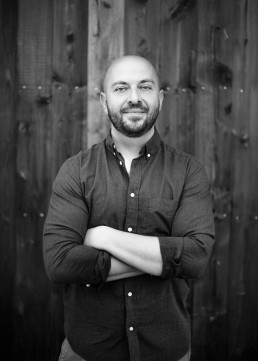 Black and white portrait of a smiling man with beard and shaved head in dark shirt against a background of a weathered antique wooden door.