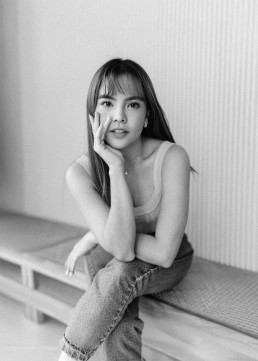 Intimate black-and-white portrait of a young Asian woman with long hair and bangs sitting on a bench, hand on cheek, pensive gaze.