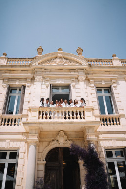 Groupe d'invités en tenues claires rassemblés sur le balcon central d'un château provençal en pierre beige avec fronton baroque, colonnes corinthiennes et balustrades ornées sous un ciel bleu