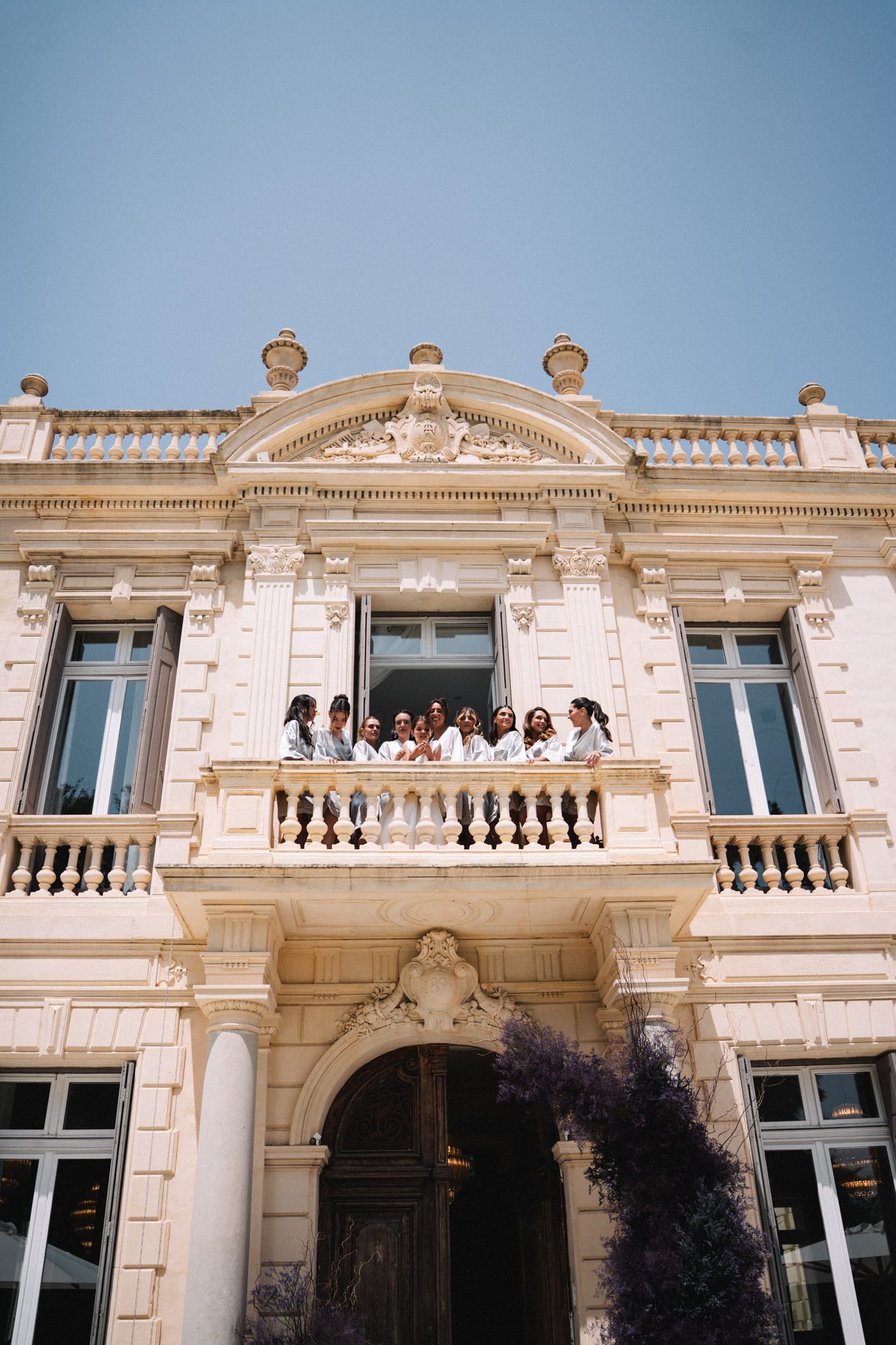 Groupe d'invités en tenues claires rassemblés sur le balcon central d'un château provençal en pierre beige avec fronton baroque, colonnes corinthiennes et balustrades ornées sous un ciel bleu