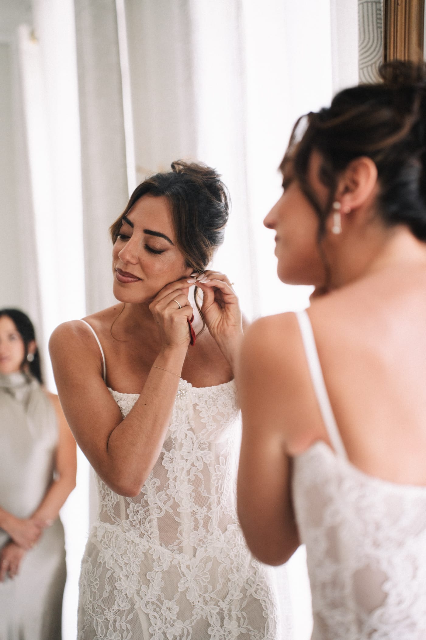 Mariée en robe bustier de dentelle blanche mettant ses boucles d'oreilles avec l'aide d'une demoiselle d'honneur, cheveux bruns relevés en chignon sophistiqué