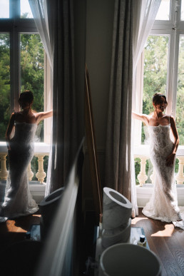 Vue miroir d'une mariée en robe blanche se tenant devant de hautes fenêtres avec rideaux verts gris, lumière naturelle douce