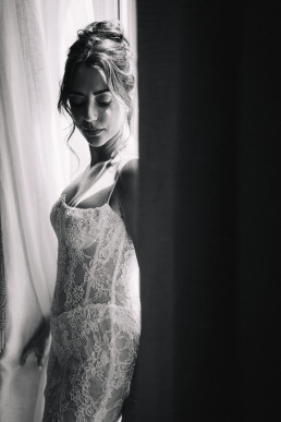 Portrait noir et blanc artistique de mariée en robe de dentelle bustier près d'une fenêtre lumineuse avec voile blanc transparent créant un effet doux et romantique