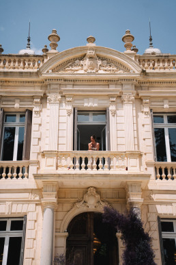 Mariée seule en robe bustier se tenant sur le balcon orné d'un château provençal avec architecture classique baroque, fronton sculpté et balustrades en pierre, ciel bleu en arrière-plan