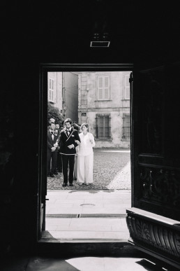 Photo noir et blanc artistique de couple de mariés vu à travers l'embrasure d'une grande porte ouverte d'un château provençal, cour pavée ensoleillée en arrière-plan
