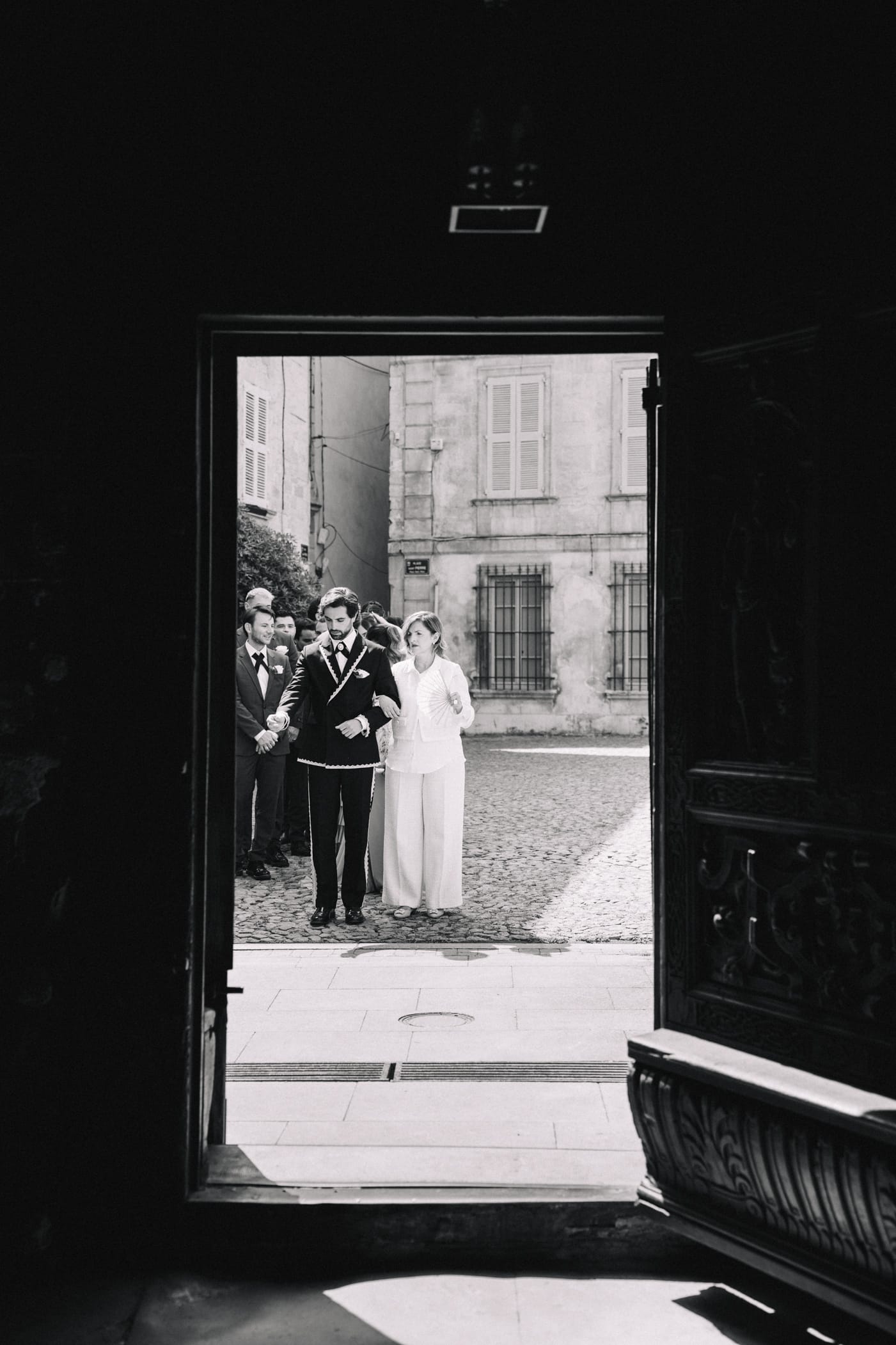 Photo noir et blanc artistique de couple de mariés vu à travers l'embrasure d'une grande porte ouverte d'un château provençal, cour pavée ensoleillée en arrière-plan