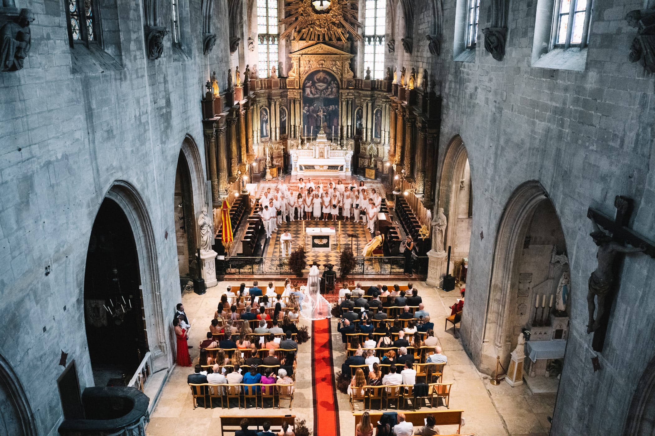 Vue aérienne plongeante de l'intérieur d'une cathédrale gothique pendant une cérémonie de mariage avec voûtes en pierre grise, autel baroque doré illuminé et invités dans les bancs