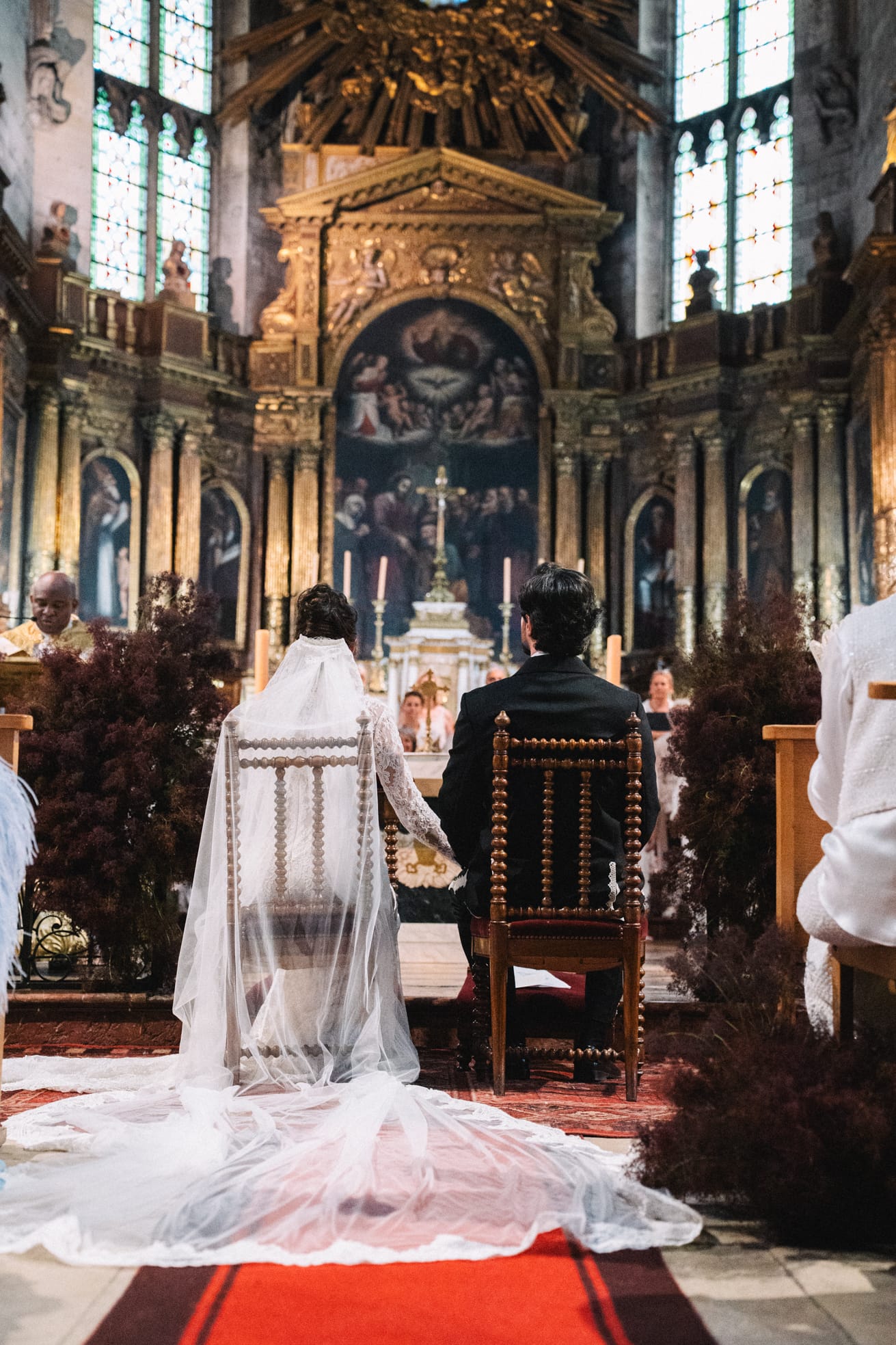 Couple de mariés de dos assis sur chaises sculptées pendant la cérémonie dans une cathédrale gothique avec autel baroque doré illuminé, long voile de dentelle blanc étalé sur tapis rouge