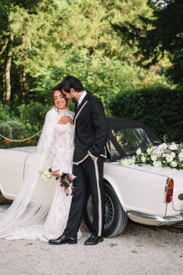 Couple de mariés riant de bonheur appuyés contre une voiture décapotable blanche vintage avec guirlande florale blanche, dans un parc verdoyant