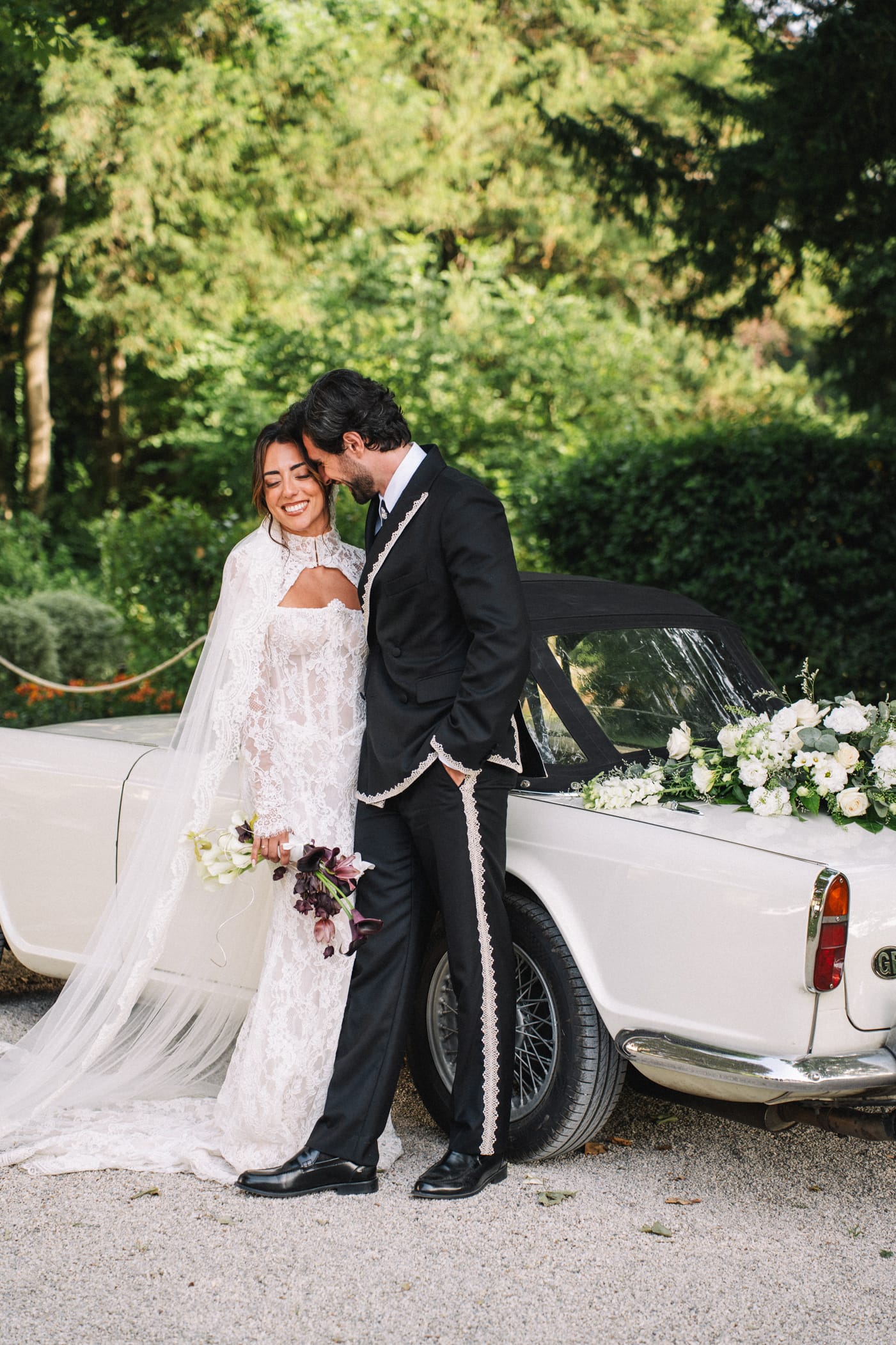 Couple de mariés riant de bonheur appuyés contre une voiture décapotable blanche vintage avec guirlande florale blanche, dans un parc verdoyant