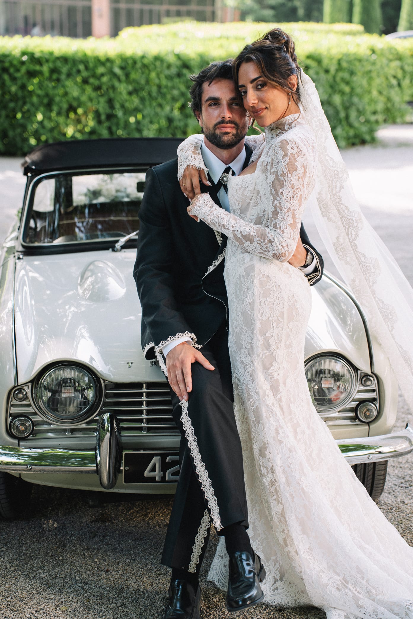 Couple de mariés posant avec voiture vintage blanche devant haie de buis taillés