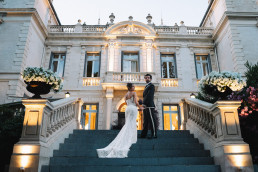 Couple de mariés posant main dans la main sur le perron illuminé d'un château provençal baroque au crépuscule, avec urnes florales blanches, éclairage architectural doré et ciel bleu