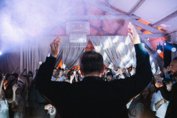 Soirée de mariage animée sous chapiteau avec invités levant les bras en dansant, machine à fumée créant une ambiance festive, drapés blancs suspendus et éclairage coloré orange et bleu