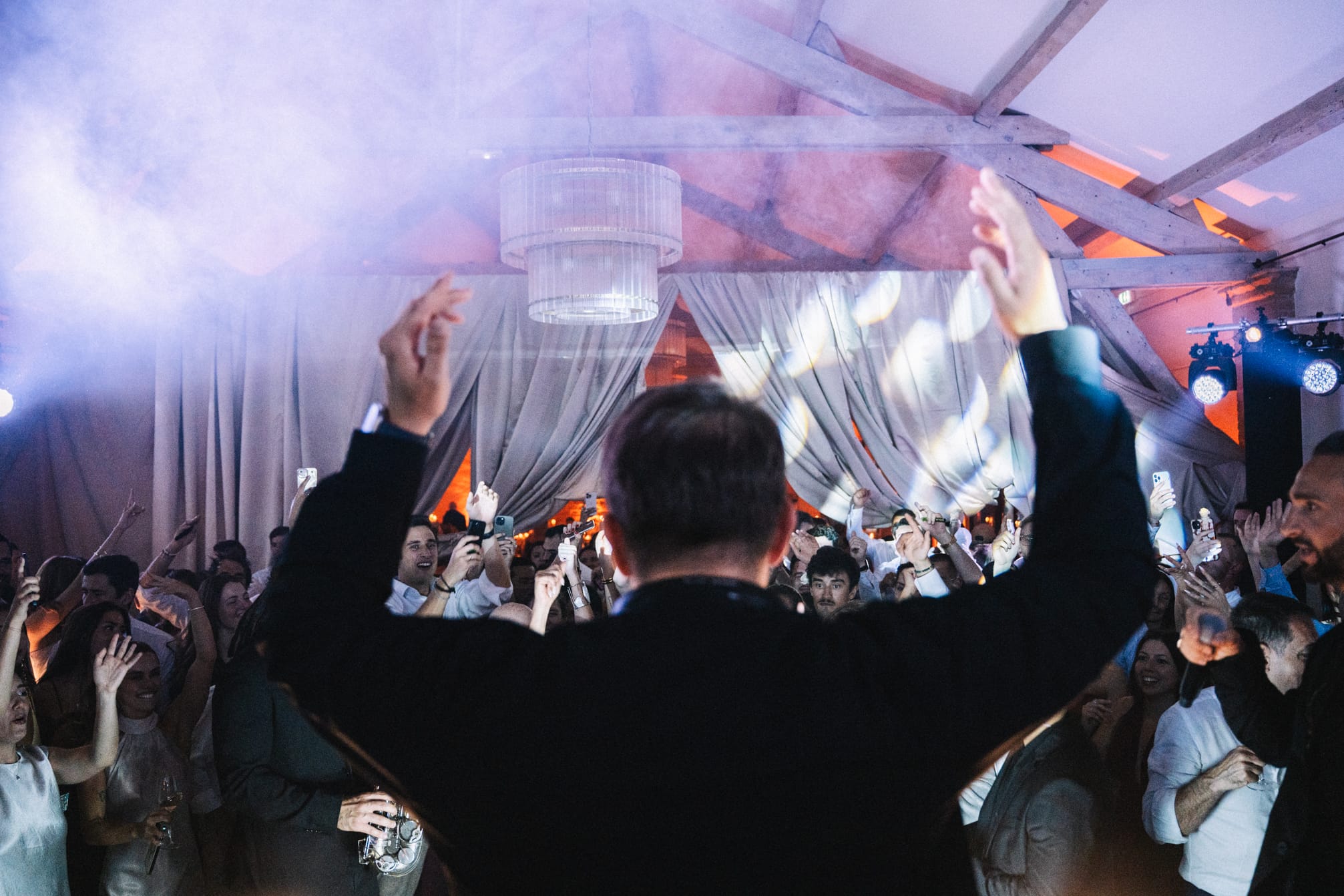 Soirée de mariage animée sous chapiteau avec invités levant les bras en dansant, machine à fumée créant une ambiance festive, drapés blancs suspendus et éclairage coloré orange et bleu