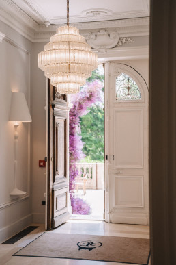 Hall d'entrée élégant d'un château provençal avec lustre en cristal à étages, moulures blanches au plafond, porte sculptée ouverte révélant un jardin avec guirlande de fleurs mauves