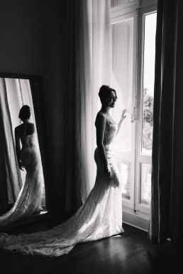 Artistic black-and-white portrait of a bride in a lace gown reflected in a large mirror near a bright window with green curtains, elegant symmetrical composition.