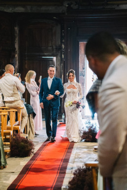The bride and groom enter a Provençal church built of ancient stone, the groom in the foreground on the right filming, the couple advancing on a red carpet with guests standing on either side.