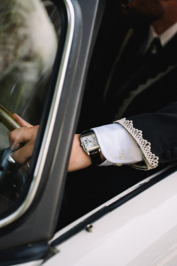 Close-up of groom's arm in black suit with elegant watch and embroidered white cuff, holding the handle of a vintage car door.