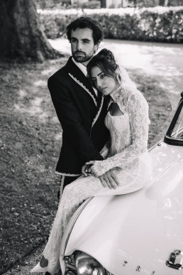 Black and white portrait of an embracing bride and groom leaning against the hood of a vintage white convertible in a lush green park.